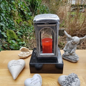 Puede incluir: Una linterna de metal plateado con una ventana de vidrio y una vela roja en su interior. La linterna está sentada sobre una base de granito negro. Hay varias piedras decorativas en forma de corazón y figuras de ángeles alrededor de la linterna.