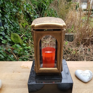 May include: A gold-coloured metal lantern with a glass window and a red candle inside. The lantern is sitting on a black granite base. There are two white heart-shaped stones with text on them in the foreground. The text on the stones reads "Wir vermissen Dich" and "In Liebe".