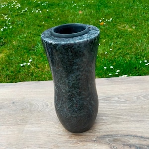 May include: A dark grey granite vase with a flared top and a curved body. The vase has a smooth, polished finish and sits on a wooden surface. The background features green grass and small white flowers.