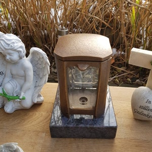 Pode incluir: Uma lanterna de exterior de cor bronze com formato retangular e painéis de vidro. A lanterna está sobre uma base de pedra cinza escuro. Uma estátua de anjo branco é visível ao fundo.