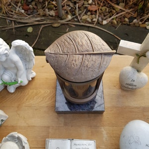 Può includere: Una lanterna commemorativa color bronzo con una parte superiore a forma di foglia, posta su una base grigio scuro. La lanterna ha un involucro di vetro. Sono visibili altri oggetti commemorativi.