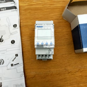 May include: A white digital timer with a black display and buttons. The timer is labeled "theben" and has a 24-hour clock face. The timer is connected to a white electrical outlet.