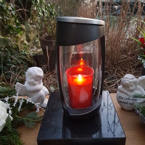Könnte beinhalten: Eine schwarze und silberne Outdoor-Kerzenlaterne mit einer roten Kerze im Inneren. Die Laterne steht auf einem schwarzen rechteckigen Sockel, umgeben von weißen Engelsstatuen und Blumengestecken. Der Hintergrund zeigt Grün.