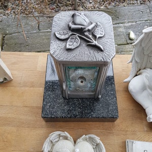 Könnte beinhalten: Eine silberfarbene Gedenklaterne mit einem Rosenmotiv auf der Oberseite. Die Laterne hat Glasscheiben und steht auf einem schwarzen Granitsockel. Weitere Gedenkgegenstände, darunter ein herzförmiger Stein mit Tauben und Text, sind ebenfalls sichtbar.