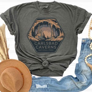 May include: Dark grey t-shirt with a hexagonal graphic of Carlsbad Caverns National Park. The graphic shows a cave interior with stalactites and stalagmites, and the text "CARLSBAD CAVERNS NATIONAL PARK". The shirt is styled with a brown hat and blue jeans.