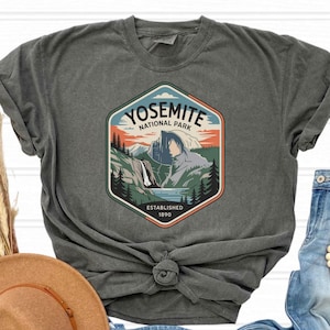 May include: Dark gray t-shirt featuring a Yosemite National Park graphic. The design includes a mountain landscape with a waterfall, trees, and the text "Yosemite National Park Established 1890". The shirt is a casual wear item.