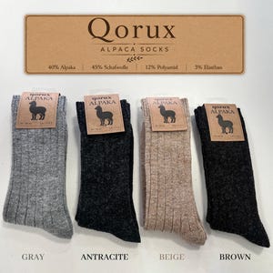 May include: Four pairs of ribbed socks in gray, anthracite, beige, and brown. Each pair features a cardboard tag with a black alpaca silhouette and the text Qorux Alpaka. The header card lists the material composition as 40% Alpaca, 45% Schafwolle, 12% Polyamid, and 3% Elasthan.