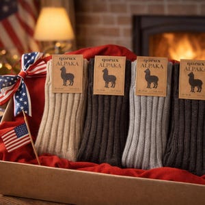 May include: A gift box filled with four pairs of ribbed alpaca wool socks in shades of beige, gray, and brown. Each pair has a tag with an alpaca logo and the brand name. The box is decorated with an American flag bow and small flags.