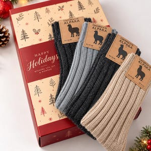 May include: Four pairs of ribbed socks in charcoal, light gray, dark gray, and beige, each with a Qorux Alpaka tag labeled EU 43-46 and US 10-12. The socks are arranged on a rectangular gift box featuring a holiday tree pattern and the text Happy Holidays Wishing You Joy & Wonder.