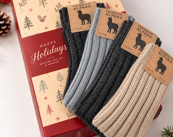 May include: Four pairs of ribbed socks in charcoal, light gray, dark gray, and beige, each with a Qorux Alpaka tag labeled EU 43-46 and US 10-12. The socks are arranged on a rectangular gift box featuring a holiday tree pattern and the text Happy Holidays Wishing You Joy & Wonder.