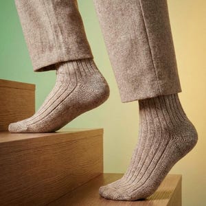 May include: A pair of light brown, ribbed knit socks worn with light brown trousers. The socks feature a vertical ribbed texture and a reinforced heel construction, extending to the mid-calf area, providing a simple and functional design.