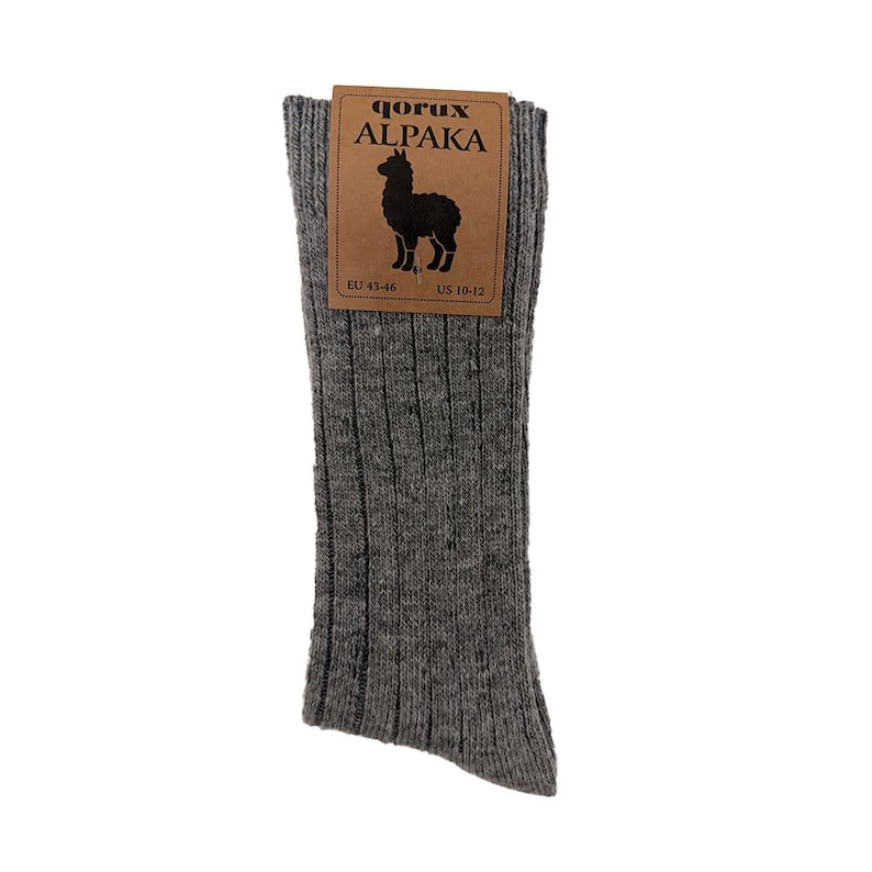 May include: A pair of grey ribbed socks with a brown paper label featuring the text QORUX ALPAKA, a silhouette of an alpaca, and size information including EU 43-46 and US 10-12. The socks have a textured, heathered appearance with vertical ribbing.