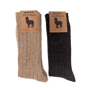 May include: A pair of ribbed socks, one light tan and one dark charcoal, each with a brown cardboard tag featuring the text QORUX ALPAKA, an illustration of an alpaca, and size information indicating EU 39-42 and US 7.5-9.5 for each pair.
