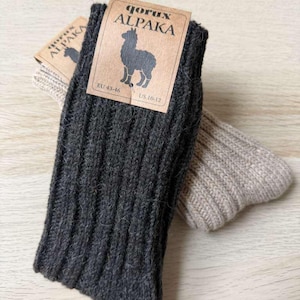 Può includere: Due paia di calzini in lana di alpaca. Un paio è grigio scuro, l'altro è grigio chiaro. Ogni paio ha una trama a coste e un'etichetta marrone con la scritta "gorux ALPAKA" e un'illustrazione di alpaca. Le taglie dei calzini sono EU 43-46.