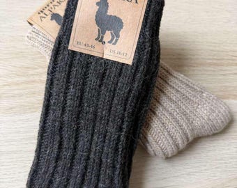 Puede incluir: Un par de calcetines de punto acanalado, uno gris carbón oscuro y otro beige claro, con una etiqueta de papel marrón que muestra el texto QORUX ALPAKA, una ilustración de una alpaca y las tallas EU 43-46 y US 10-12 impresas en la etiqueta.
