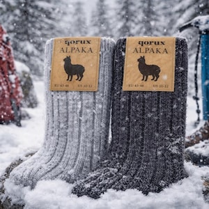 May include: Two pairs of alpaca wool socks, one light gray and one dark gray, are displayed in a snowy outdoor setting. Each sock has a tag with the text "qorux ALPAKA" and a silhouette of an alpaca. The socks are EU size 43-46 and US size 10-12.