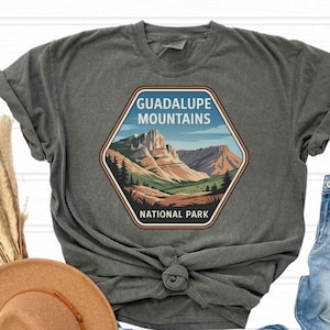 May include: Dark gray t-shirt featuring a hexagonal graphic of Guadalupe Mountains National Park. The graphic includes the text "GUADALUPE MOUNTAINS" and "NATIONAL PARK" with a mountain landscape in the center. The shirt is made of a soft material.