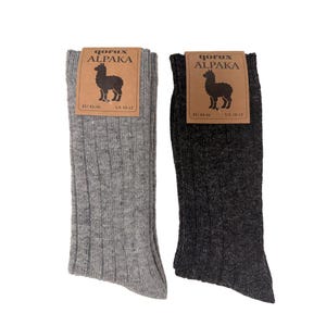May include: Two pairs of ribbed socks, one light gray and one dark charcoal gray. Each pair features a brown cardboard label with the text qorux ALPAKA, an illustration of an alpaca, and the size information EU 43-46 and US 10-12 printed on it.