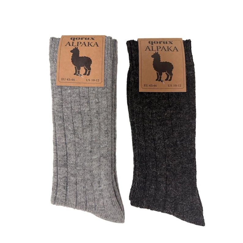 May include: Two pairs of ribbed socks, one light gray and one dark charcoal gray. Each pair features a brown cardboard label with the text qorux ALPAKA, an illustration of an alpaca, and the size information EU 43-46 and US 10-12 printed on it.