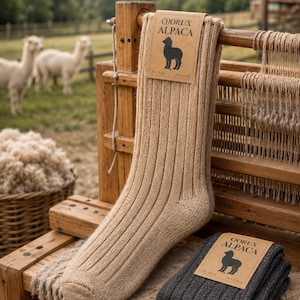May include: A pair of ribbed knit socks in tan and dark gray, each featuring a tan paper label with the text QORUX ALPACA, an alpaca silhouette, and size markings EU 38-42 and US 7.5-10.5. The tan sock is upright, while the dark gray pair is folded.