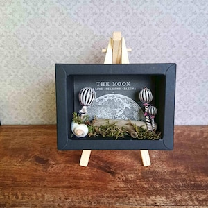 Könnte beinhalten: Ein Miniatur-Diorama in einem schwarzen Schattenkasten, das eine Mondszene darstellt. Das Diorama umfasst einen strukturierten Mond, gestreifte Heißluftballons und eine Muschel. Der Text "THE MOON" ist sichtbar.