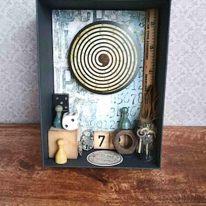 Könnte beinhalten: Eine dekorative Schattenbox mit einem Spiraldesign, Würfeln, einem Lineal und anderen Vintage-Objekten. Die Farbpalette umfasst Brauntöne, Beige und Blau. Die Zahl "7" ist sichtbar, zusammen mit einer kleinen Glasflasche und einem Schlüssel.