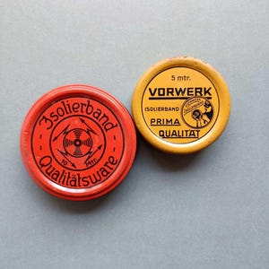 Könnte beinhalten: Zwei runde Vintage-Dosen. Eine ist rot mit schwarzem Text und einer Grafik, die andere ist gelb mit schwarzem Text und einem Logo. Auf der roten Dose steht "3 Isolierband Qualitätsware" und auf der gelben Dose steht "VORWERK Isolierband PRIMA QUALITAT".