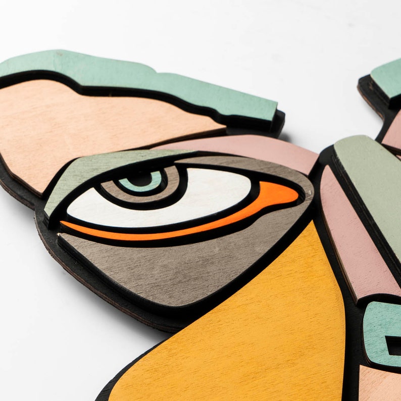 May include: A close-up of a wooden wall art piece featuring a stylized eye with orange, brown, and white details. The eye is part of a larger abstract design with shades of pink, yellow, and green.