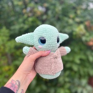Pu&ograve; includere: Un peluche all'uncinetto raffigurante un personaggio con grandi occhi e orecchie a punta. Il giocattolo &egrave; principalmente verde chiaro con una giacca marrone. Il giocattolo &egrave; tenuto in una mano con un tatuaggio.