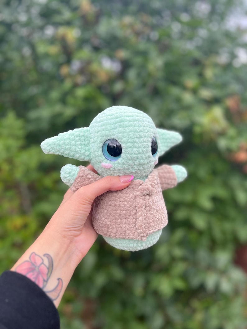 Pu&ograve; includere: Un peluche all'uncinetto raffigurante un personaggio con grandi occhi e orecchie a punta. Il giocattolo &egrave; principalmente verde chiaro con una giacca marrone. Il giocattolo &egrave; tenuto in una mano con un tatuaggio.