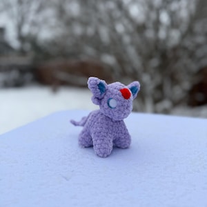 Pu&ograve; includere: Una piccola creatura all'uncinetto color lavanda con orecchie blu e un segno rosso a forma di cuore sulla fronte. Il giocattolo fatto a mano ha una lunga coda ed &egrave; seduto su una superficie innevata. Lo sfondo &egrave; sfocato, con rami di alberi spogli visibili.