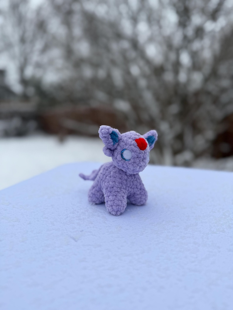 Pu&ograve; includere: Una piccola creatura all'uncinetto color lavanda con orecchie blu e un segno rosso a forma di cuore sulla fronte. Il giocattolo fatto a mano ha una lunga coda ed &egrave; seduto su una superficie innevata. Lo sfondo &egrave; sfocato, con rami di alberi spogli visibili.