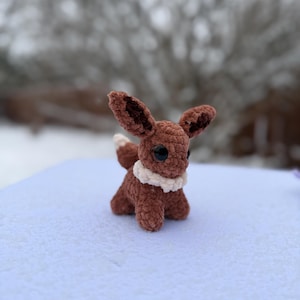 Pu&ograve; includere: Un peluche Eevee marrone all'uncinetto fatto a mano con grandi occhi neri, un colletto bianco e una coda soffice. Il peluche &egrave; ambientato su uno sfondo innevato, mostrando la sua lavorazione dettagliata e la sua morbida consistenza. Eevee misura circa 13 cm di altezza.