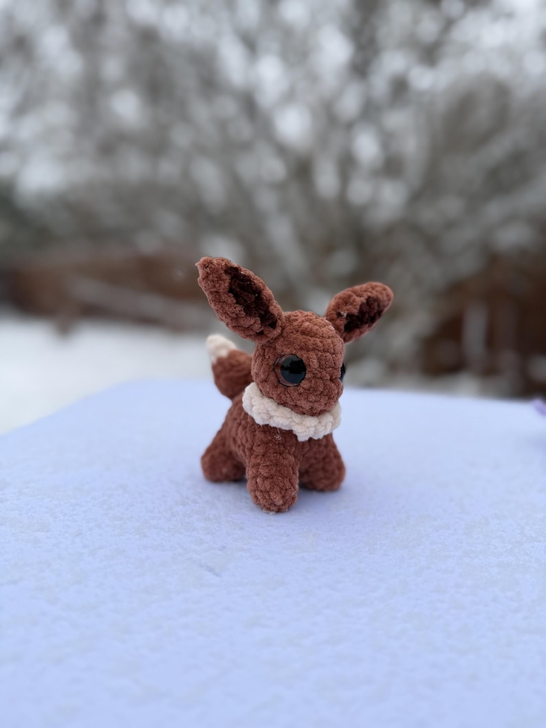 Pu&ograve; includere: Un peluche Eevee marrone all'uncinetto fatto a mano con grandi occhi neri, un colletto bianco e una coda soffice. Il peluche &egrave; ambientato su uno sfondo innevato, mostrando la sua lavorazione dettagliata e la sua morbida consistenza. Eevee misura circa 13 cm di altezza.