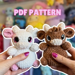 May include: Two crocheted cow plushies, one white with pink accents and the other brown. Both have large, sparkly eyes and the text "PDF PATTERN" in pink above them. These handmade toys are soft and cuddly.