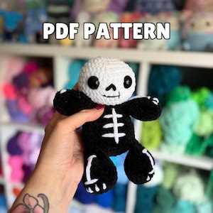 Peut inclure: Une poupée squelette au crochet avec une tête de crâne blanche aux yeux noirs et un corps noir avec des détails d'os de cage thoracique et de membres blancs. Le texte PDF PATTERN est affiché en lettres blanches sur un fond flou.