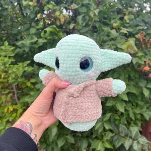 Pu&ograve; includere: Un peluche all'uncinetto raffigurante un personaggio di una popolare serie di fantascienza. Il giocattolo &egrave; principalmente verde chiaro con grandi occhi neri e blu e un abito color cachi. Il giocattolo ha grandi orecchie e braccia piccole.