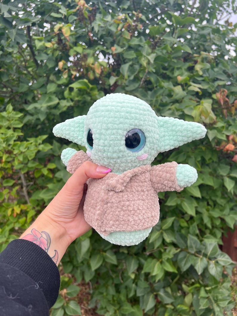 Pu&ograve; includere: Un peluche all'uncinetto raffigurante un personaggio di una popolare serie di fantascienza. Il giocattolo &egrave; principalmente verde chiaro con grandi occhi neri e blu e un abito color cachi. Il giocattolo ha grandi orecchie e braccia piccole.