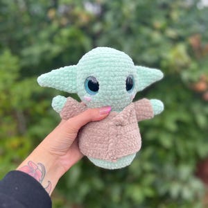 Pu&ograve; includere: Bambola all'uncinetto raffigurante un personaggio popolare, con testa e orecchie verde chiaro, grandi occhi neri con accenti rosa e un cappotto color cammello. La bambola &egrave; tenuta in mano.