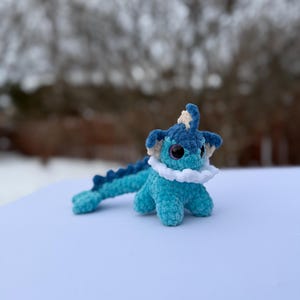 Puede incluir: Un peluche de ganchillo hecho a mano en tonos azules y blancos. El juguete tiene ojos grandes y redondos, un cuello blanco con volantes y una cola larga y segmentada. El peluche est&aacute; sobre una superficie blanca con un fondo borroso.