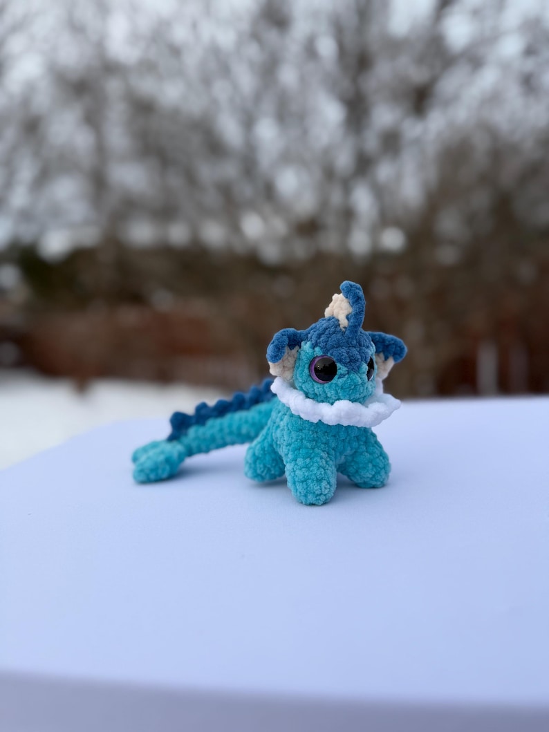 Puede incluir: Un peluche de ganchillo hecho a mano en tonos azules y blancos. El juguete tiene ojos grandes y redondos, un cuello blanco con volantes y una cola larga y segmentada. El peluche est&aacute; sobre una superficie blanca con un fondo borroso.