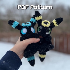 Könnte beinhalten: Zwei schwarze Eevee-Plüschtiere aus Häkelarbeit mit blauen und gelben Akzenten. Die Plüschtiere haben große, glitzernde Augen und werden von einer Hand gehalten.