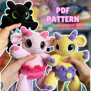 May include: A collection of handmade crochet plushies in various colors. The plushies include a black dragon with green eyes, a pink creature with a bow, and a yellow creature with purple accents. The text "PDF PATTERN" is visible.
