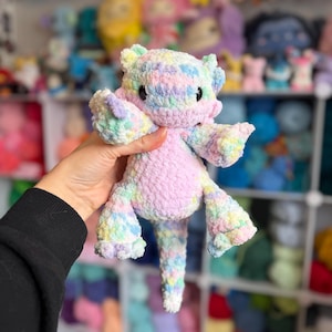 Peut inclure: Une peluche axolotl au crochet faite à la main, aux couleurs pastel. La peluche présente un corps rose clair, avec des accents pastel multicolores sur la tête, les bras et les jambes. L'axolotl a de petits yeux noirs et est tenu dans une main.