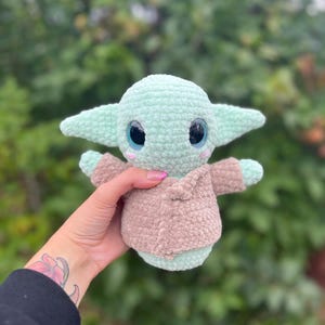 Pu&ograve; includere: Bambola all'uncinetto di un personaggio con grandi occhi, grandi orecchie e un cappotto color cammello. La bambola &egrave; verde chiaro con un cappotto color cammello e grandi occhi neri con accenti blu. La bambola &egrave; tenuta davanti a uno sfondo verde sfocato.