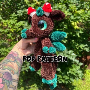 Puede incluir: Un peluche de dragón marrón tejido a ganchillo con detalles turquesa en las alas y los pies, y acentos de setas rojas y blancas en la cabeza. El dragón tiene ojos grandes y reflectantes. El texto "PDF PATTERN" se muestra en la imagen.