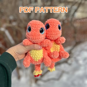 May include: Two crocheted plush toys in the shape of a dinosaur. The toys are orange with yellow accents and blue eyes. The toys are being held in a person's hand. The image includes the text "PDF PATTERN".