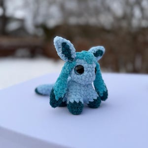 Puede incluir: Un peluche de ganchillo hecho a mano en tonos azules y turquesas. El peluche presenta ojos grandes y redondos, orejas largas y una cola. El juguete est&aacute; sobre una superficie blanca, con un fondo exterior borroso.