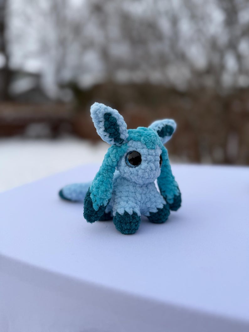Puede incluir: Un peluche de ganchillo hecho a mano en tonos azules y turquesas. El peluche presenta ojos grandes y redondos, orejas largas y una cola. El juguete est&aacute; sobre una superficie blanca, con un fondo exterior borroso.