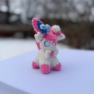 Puede incluir: Un peluche Sylveon hecho a mano con ganchillo. El peluche presenta un cuerpo blanco con detalles rosas en los pies, las orejas y la cola. Las orejas son rosas y azules, y los ojos son blancos con un centro negro. El peluche est&aacute; sobre una superficie blanca.